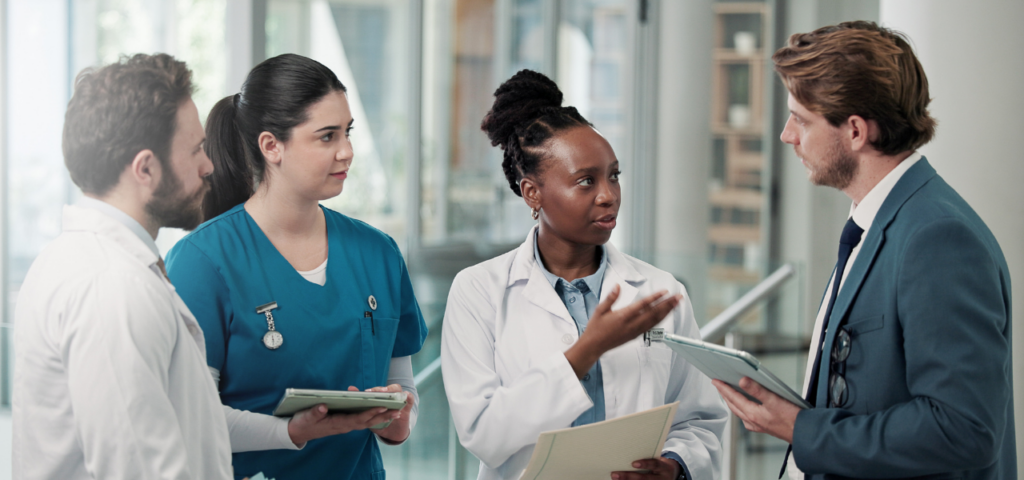 Four people talking in a medical setting. They are a mix of professions, with one wearing scrubs, one in a suit, and two in lab coats.